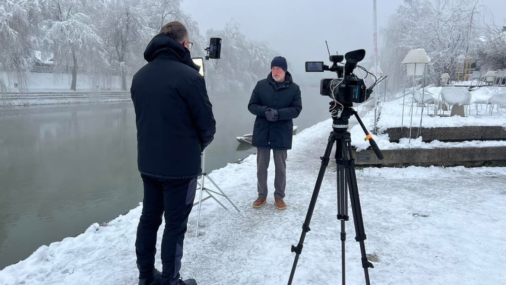 Cette image montre un homme debout au bord d’une rivière gelée, sur un sol enneigé, vêtu de noir avec de la neige en arrière-plan. Il fait face à un autre homme et à une caméra qui semble l'interviewer.
