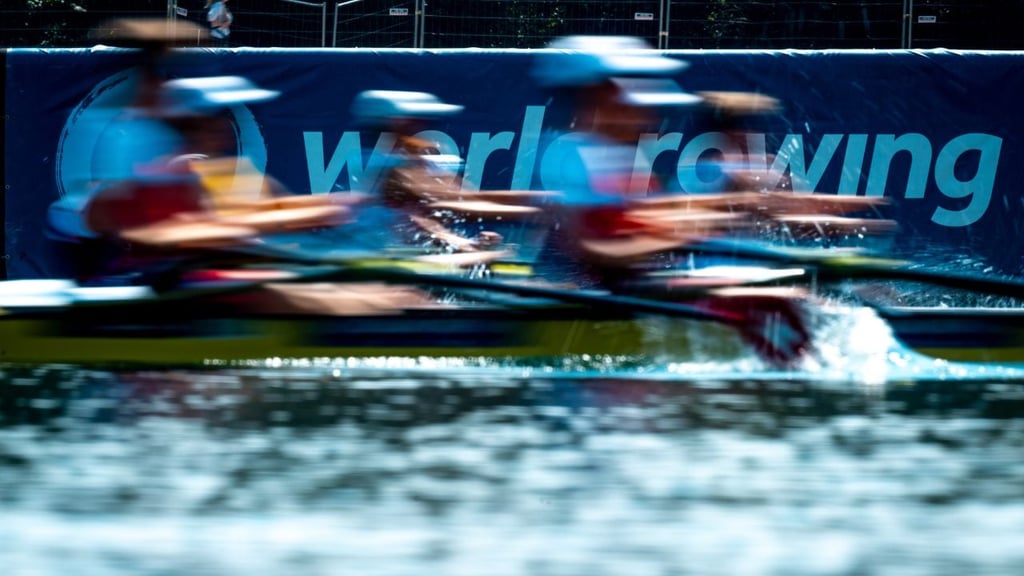 Rameurs en action sur l'eau lors d'une course, avec une bannière "World Rowing" visible à l'arrière-plan.