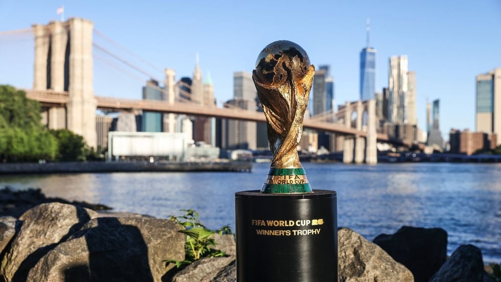 The FIFA World Cup trophy displayed outdoors on a pedestal reading ‘FIFA World Cup Winner’s Trophy,’ with the Brooklyn Bridge and Manhattan skyline in the background.