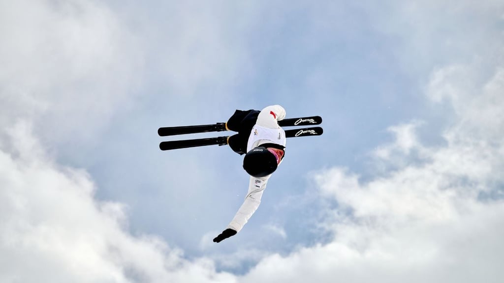 A freestyle skier performs an aerial trick high above the snow, silhouetted against a blue winter sky.