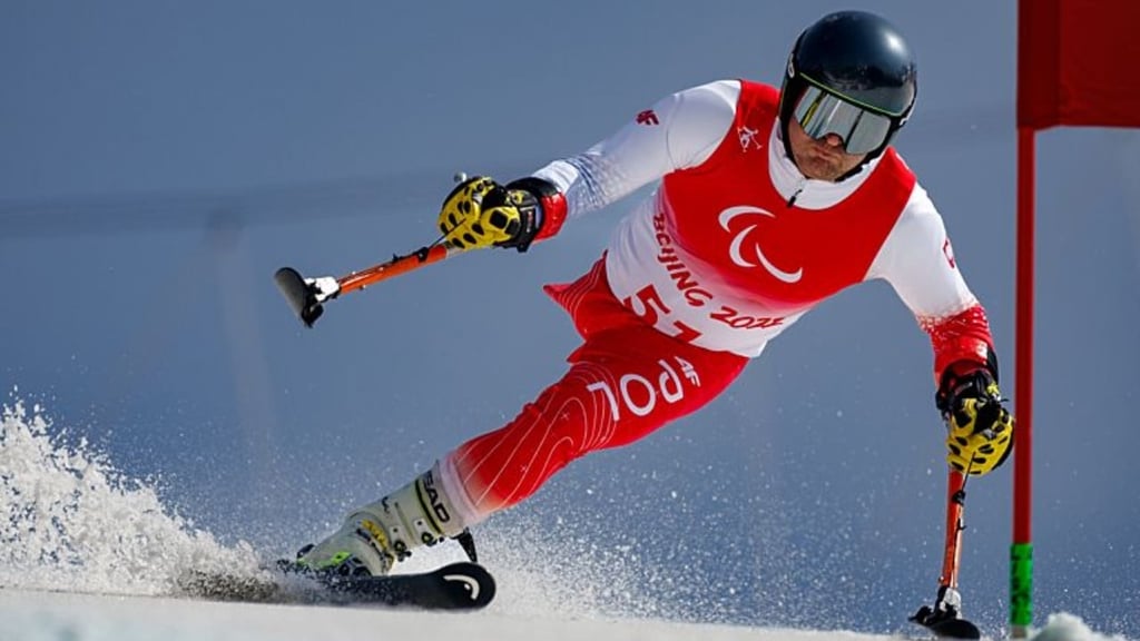 A para-alpine skier in a red and white racing suit marked “Beijing 2022” speeds down a snowy slope, leaning sharply into a turn around a slalom gate. The athlete wears a black helmet and reflective goggles, using ski poles for balance as snow sprays up from the skis.