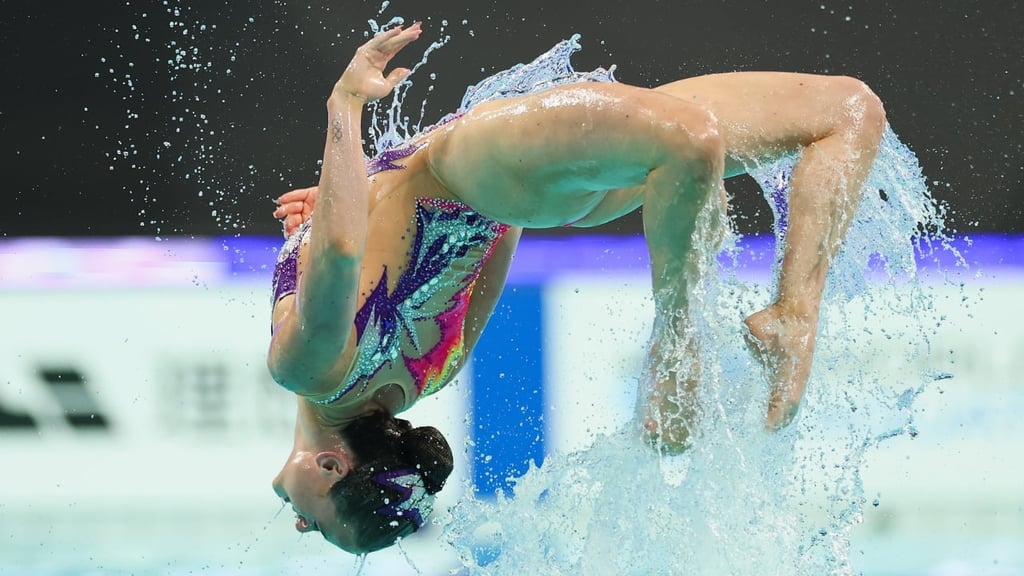 Isabelle Thorpe (Great Britain) in a colorful, patterned swimsuit launches backward out of a pool, arched in a tight backflip with water splashing dramatically around her. Her body is fully extended and curved, head tilted back toward the water, with droplets frozen midair against a blurred indoor competition backdrop.
