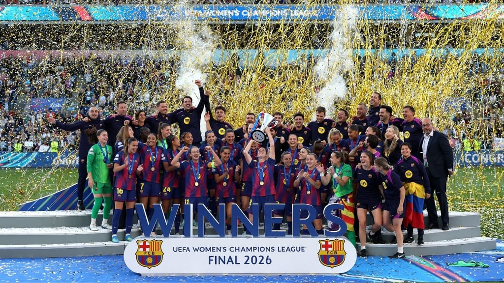 FC Barcelona Women celebrate winning the UEFA Women’s Champions League Final 2026, lifting the trophy on a podium as gold confetti rains down in a packed stadium.