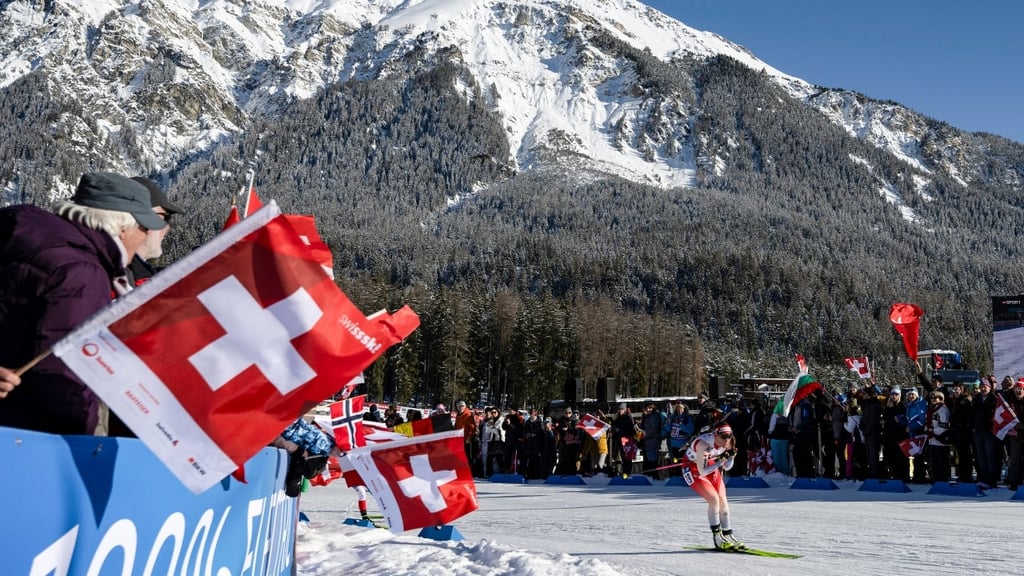 Il s'agit d'une photo de Lena Haecki-Gross sur des skis avec en arrière-plan un paysage de montagne enneigée, les fans la regardent de chaque côté alors qu'elle passe, ils tiennent des drapeaux suisses